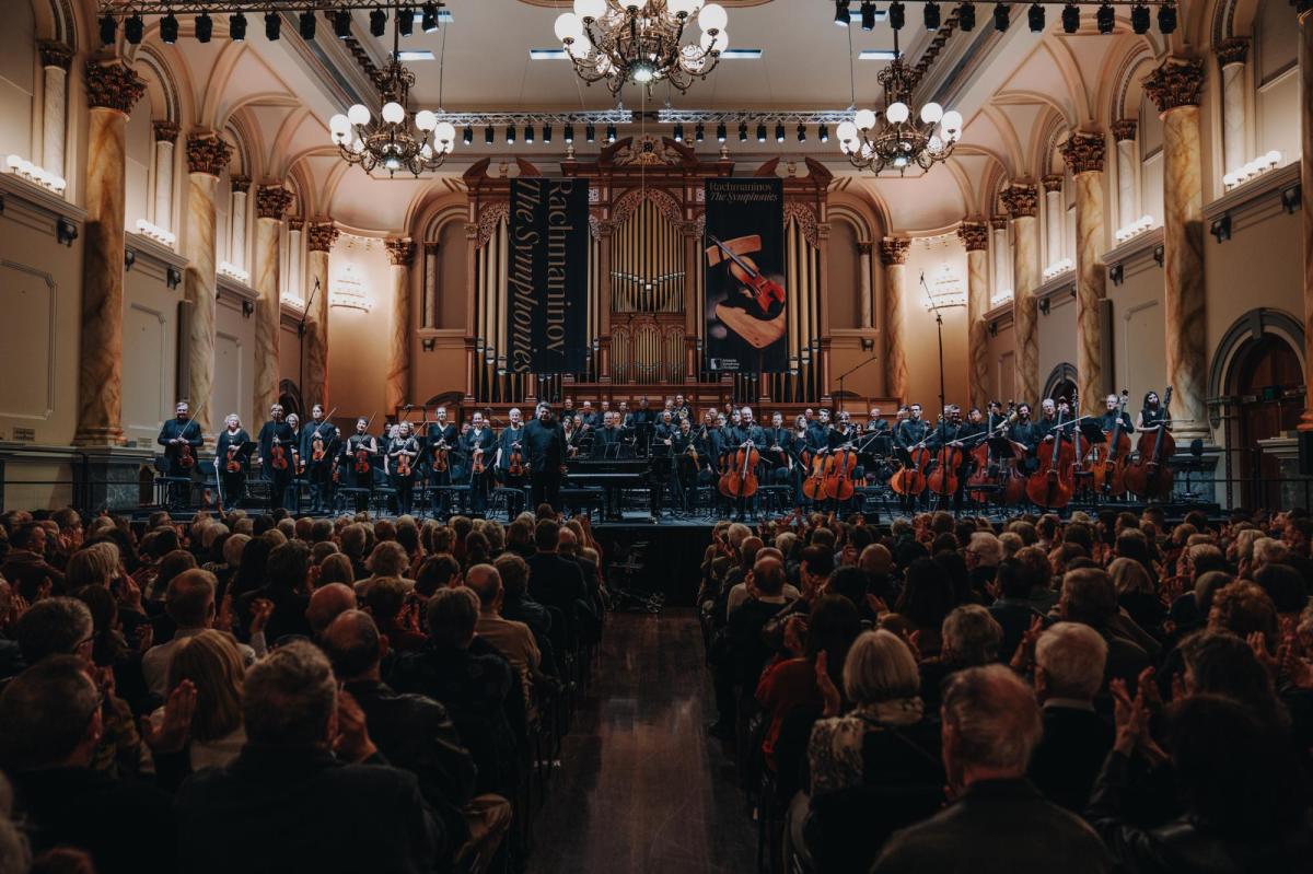 A concert hall. At the front can be seen rows of audience. Further back is the orchestra, with mostly woodwind instruments.